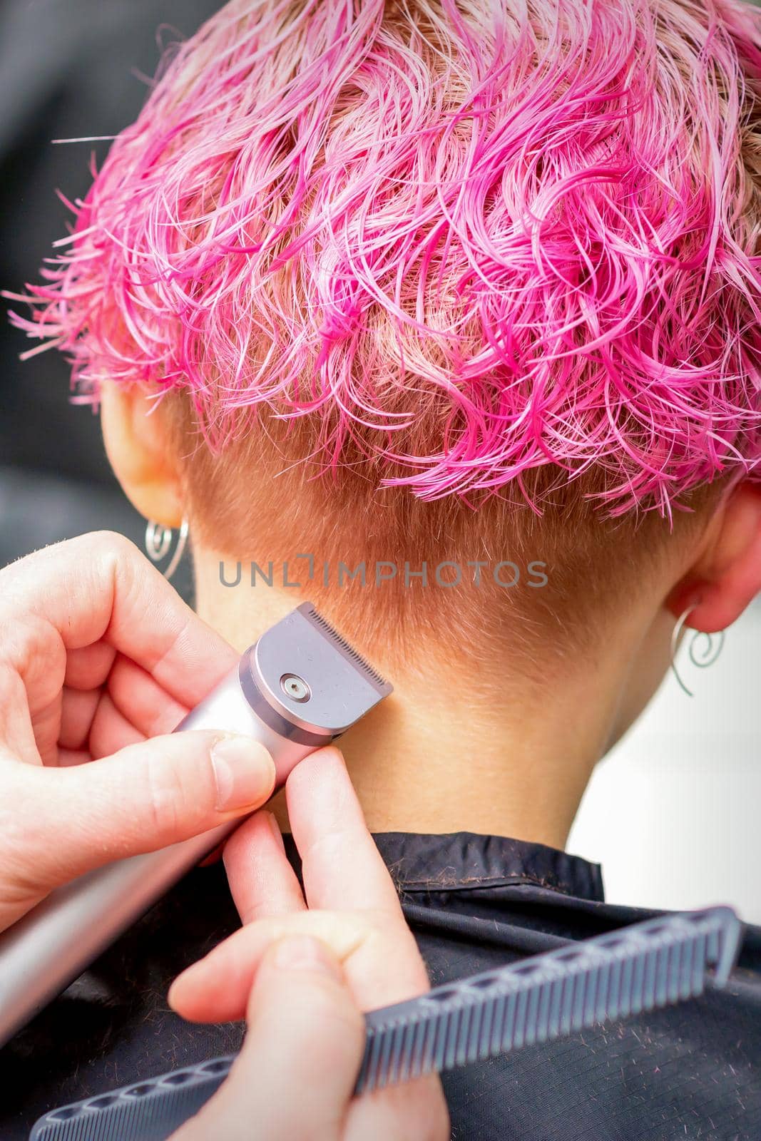 Hairdresser shaving nape and neck with electric trimmer of a young caucasian woman with short pink hair in a beauty salon. by okskukuruza