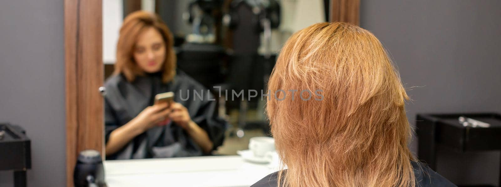 Beautiful young red hair woman using her smartphone and texting sitting in front of a mirror waiting to visit a hairdresser. by okskukuruza