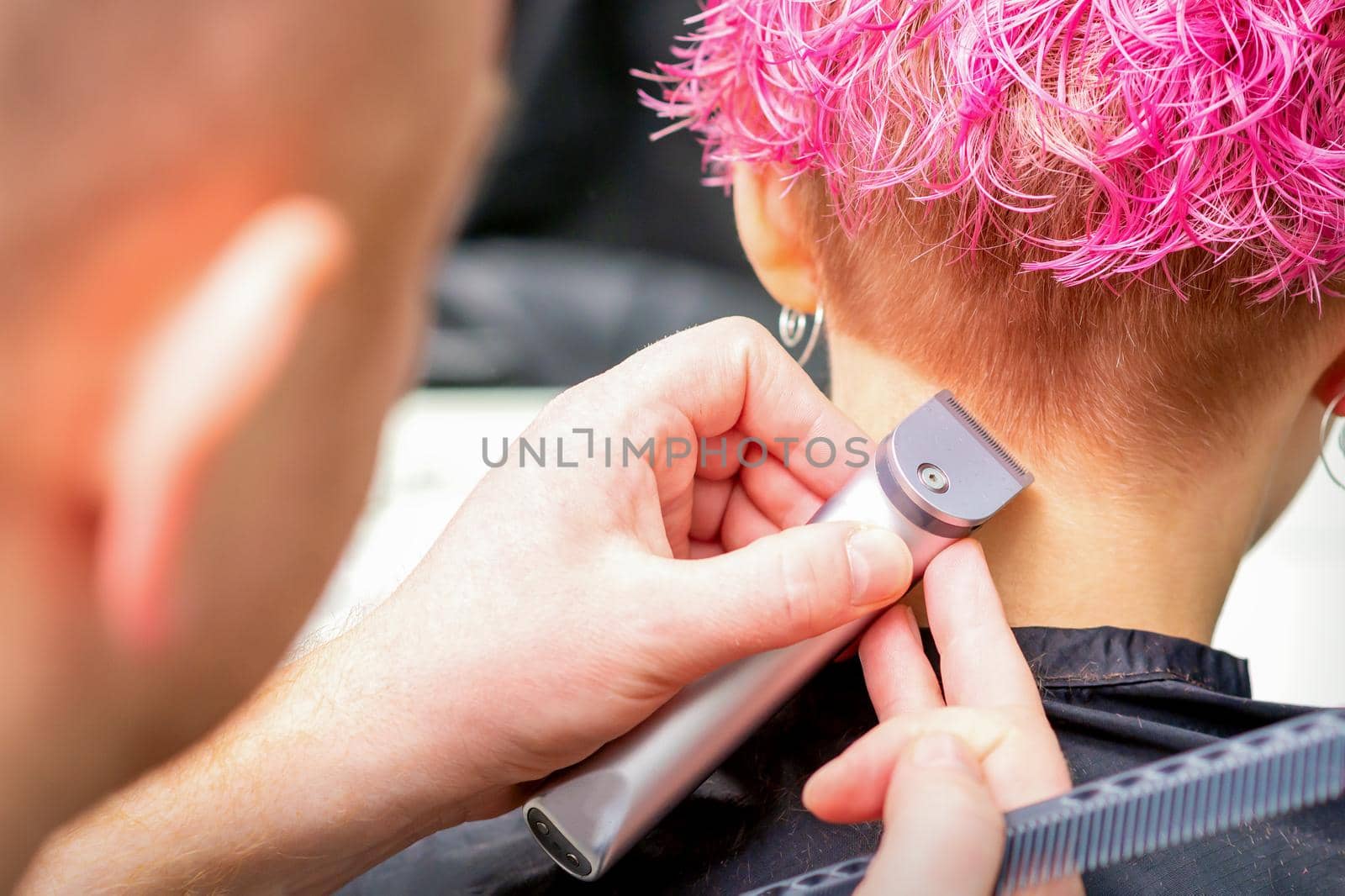 Hairdresser shaving nape and neck with electric trimmer of a young caucasian woman with short pink hair in a beauty salon