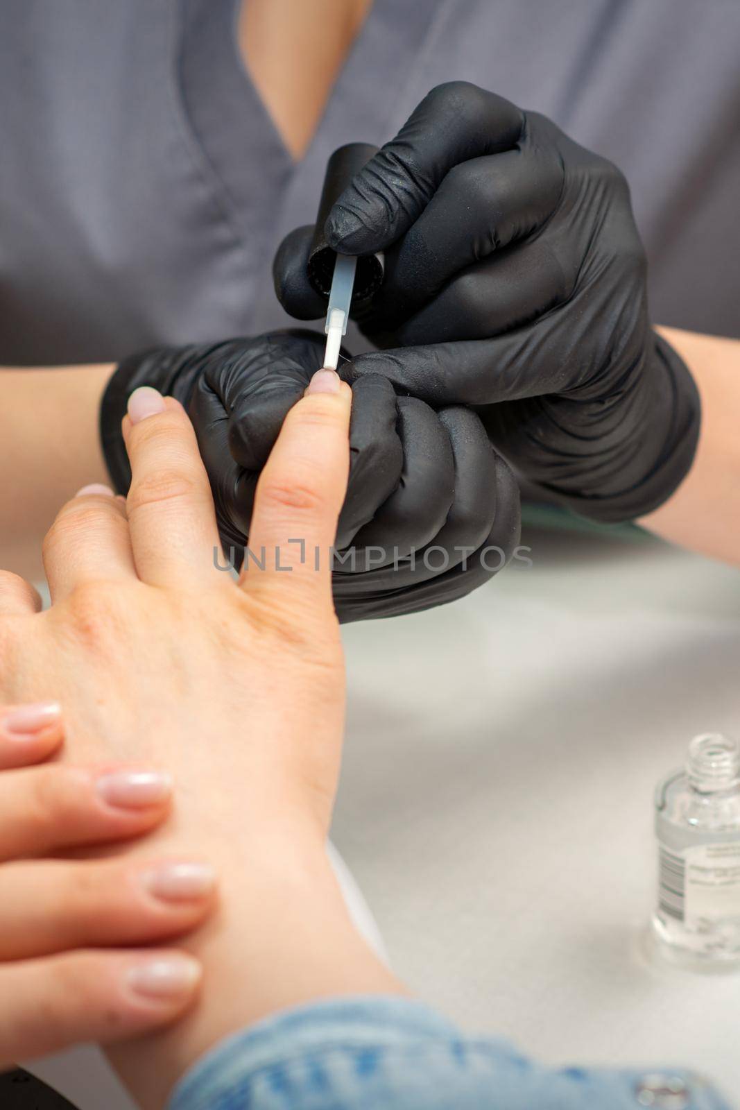 Painting female nails. Hands of manicurist in black gloves is applying transparent nail polish on female nails in a manicure salon. by okskukuruza