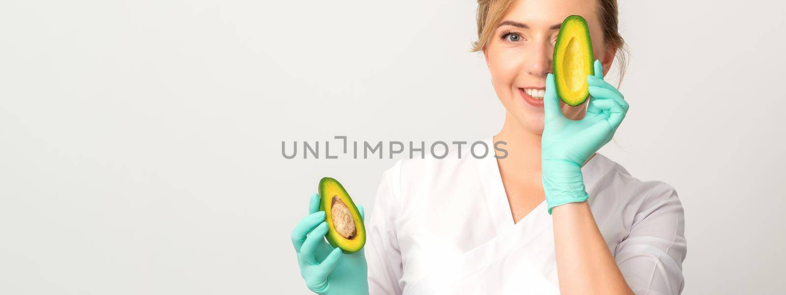 Portrait of young female nutritionist doctor with beautiful smile posing at camera hiding eye behind half avocado on white background, copy space. Benefits of proper nutrition. by okskukuruza