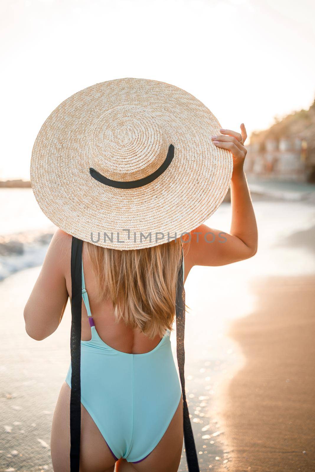 A young tanned woman in a beautiful swimsuit with a straw hat stands and rests on a tropical beach with sand and looks at the sunset and the sea. Selective focus. Vacation concept by the sea
