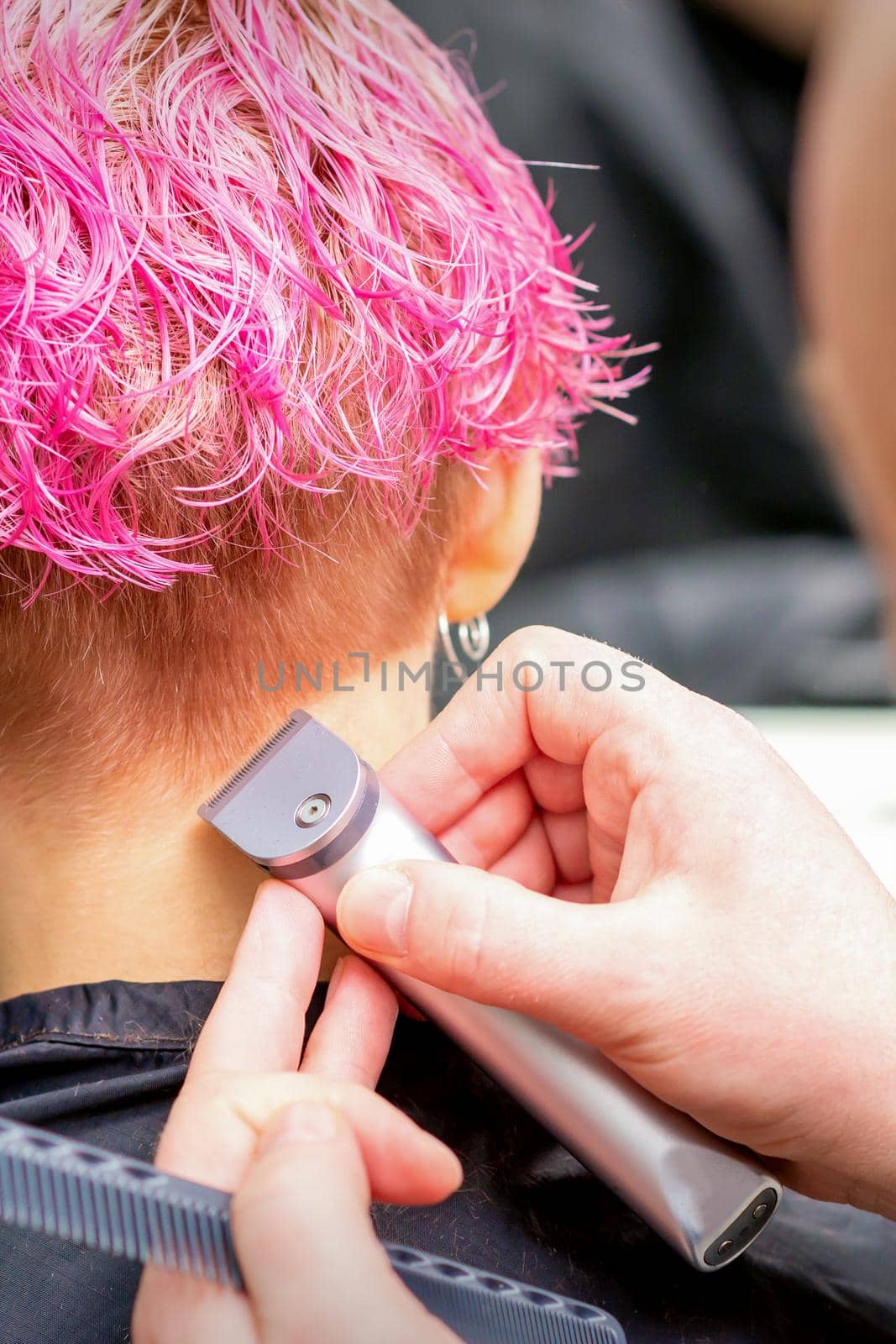 Hairdresser shaving nape and neck with electric trimmer of a young caucasian woman with short pink hair in a beauty salon. by okskukuruza