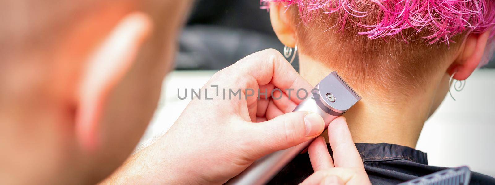 Hairdresser shaving nape and neck with electric trimmer of a young caucasian woman with short pink hair in a beauty salon. by okskukuruza