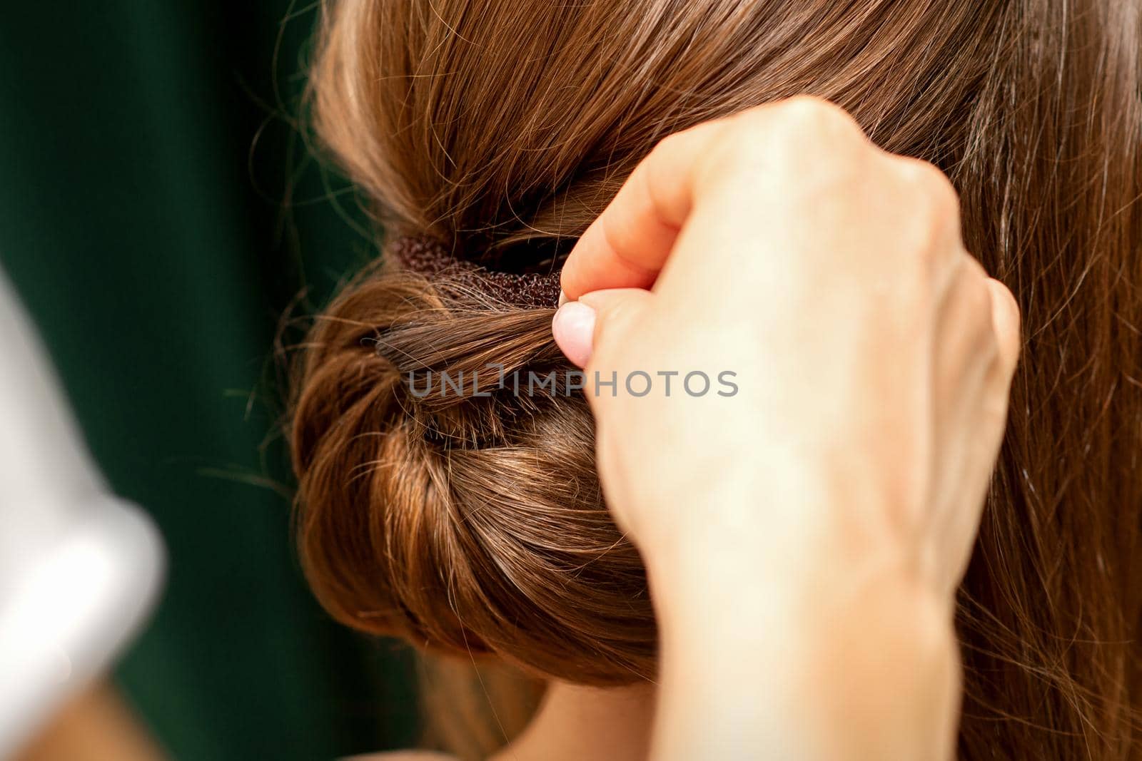 Hands of hairdresser making french twist hairstyle of an unrecognizable young brunette woman in a beauty salon, back view, close up. by okskukuruza