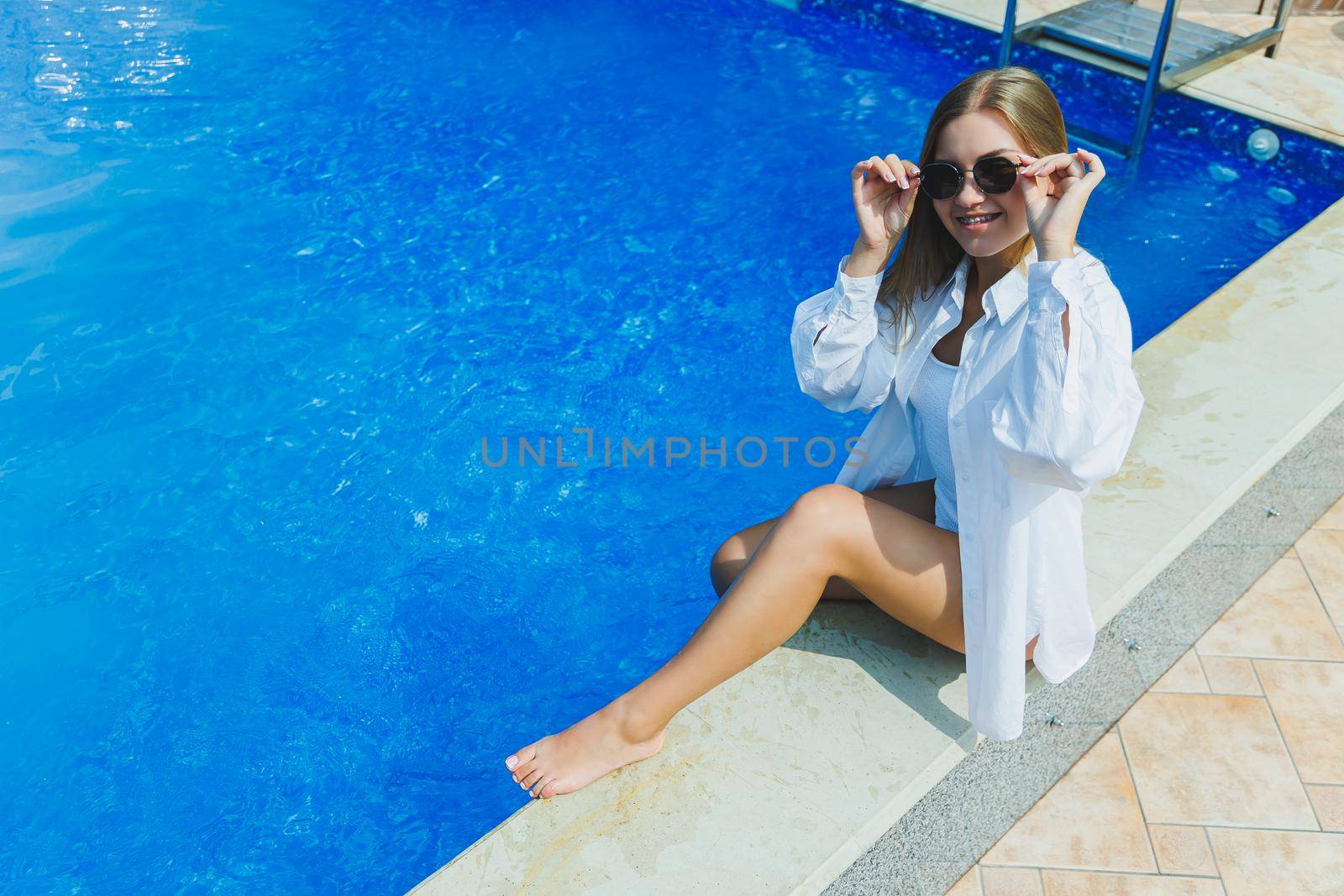 A cheerful young woman in a white swimsuit and a white shirt is sitting by the pool. A woman in sunglasses and a swimsuit. Summer vacation by the pool. Tanned girl