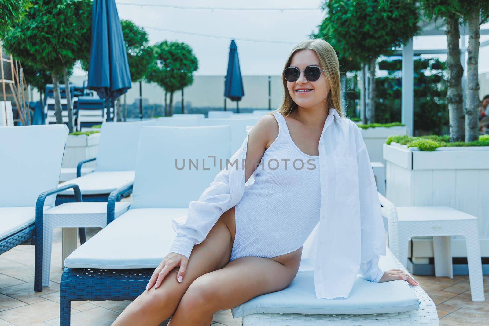 Slim woman in white swimsuit, sunglasses lying on deckchair and sunbathing by pool during summer vacation