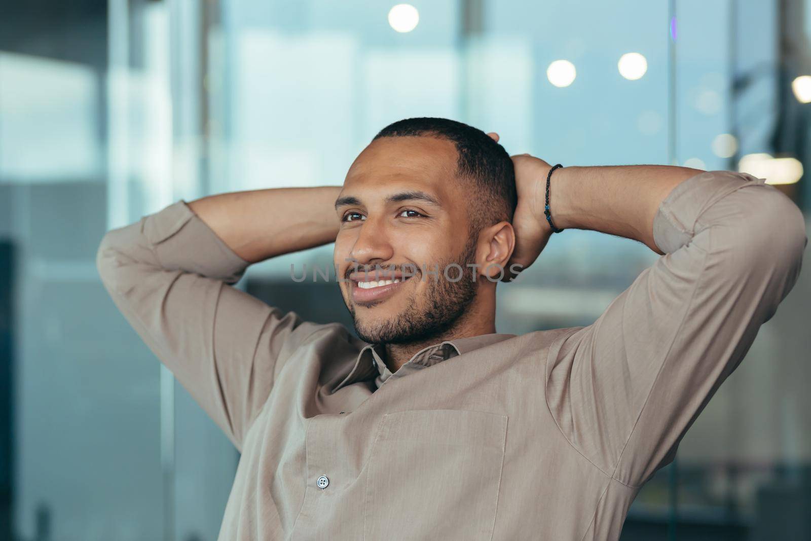 Close up photo of mixed race man relaxing in office with hands behind head looking out window and smiling, businessman daydreaming inside modern office building, successful investor happy with work
