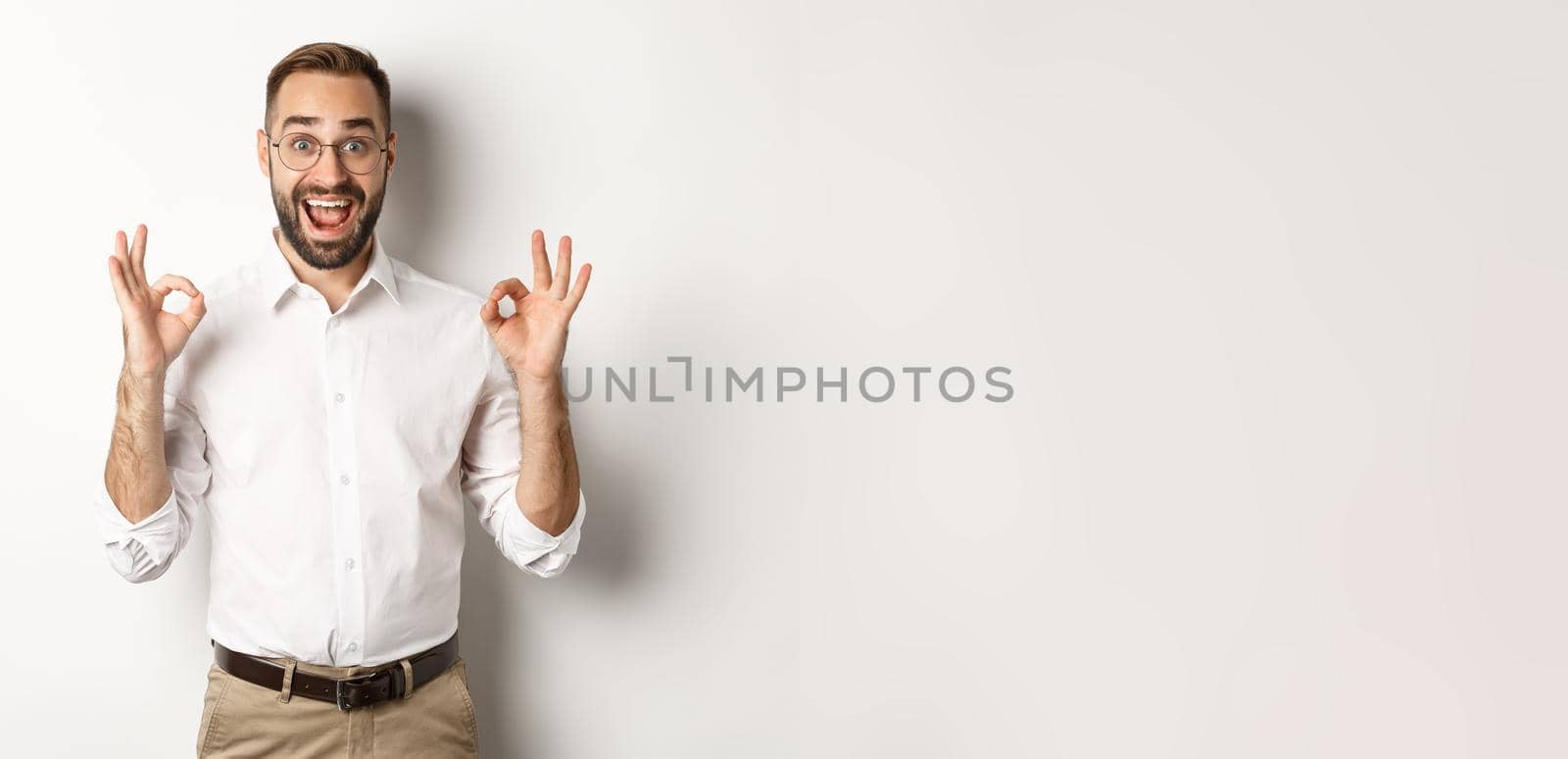 Amazed entrepreneur showing okay sign and looking happy, satisfied with product, standing over white background. Copy space