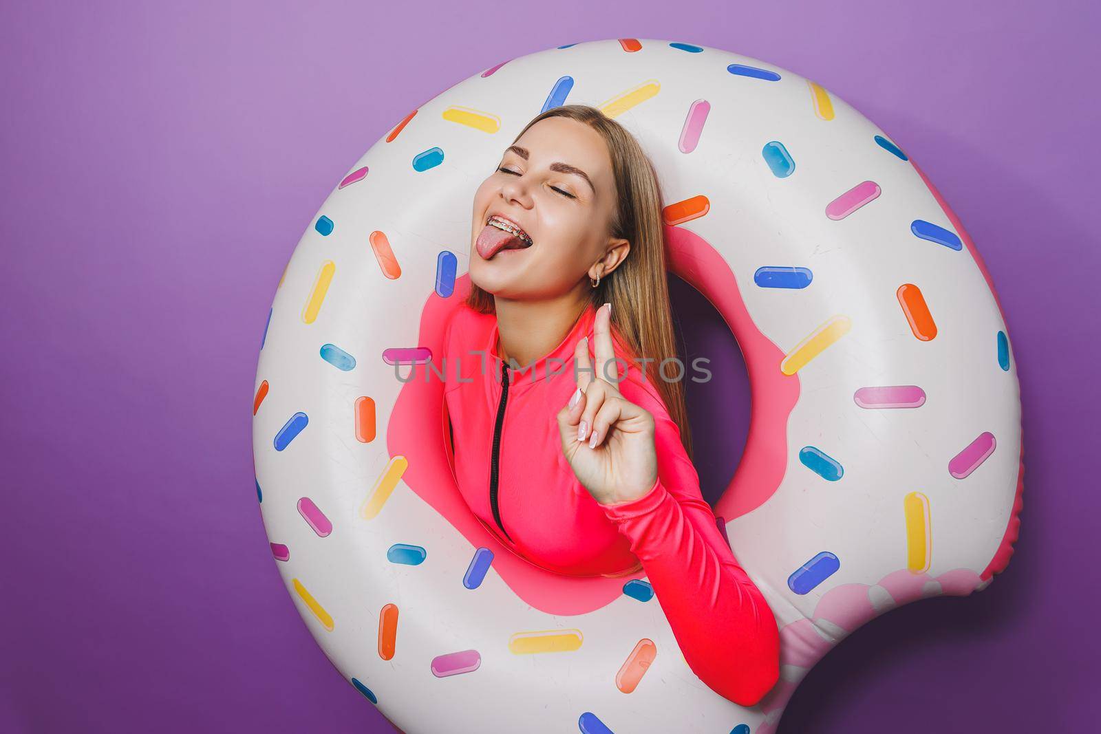 Cheerful slender woman with long hair holding an inflatable ring on a purple background. Attractive girl in a pink swimsuit by Dmitrytph