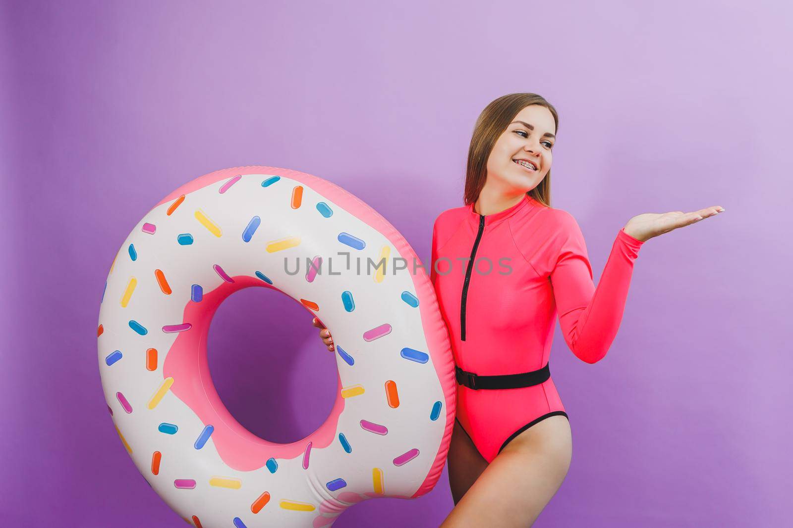 Slim young woman in a stylish pink swimsuit with a donut inflatable ring on a plain background.
