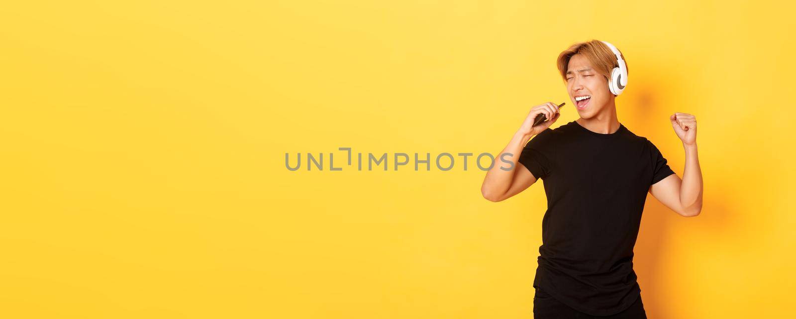 Carefree handsome asian guy in headphones, playing karaoke app, singing into mobile phone microphone, standing yellow background.