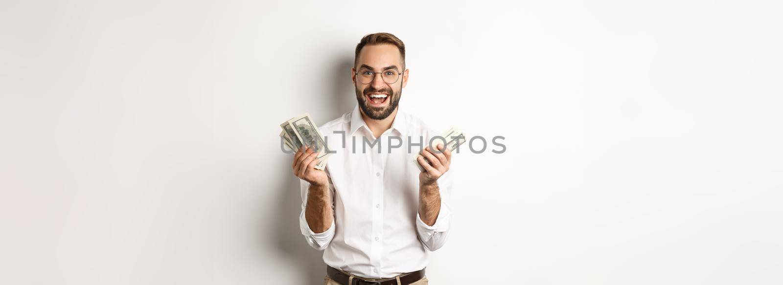 Handsome successful business man counting money, rejoicing and smiling, standing over white background.