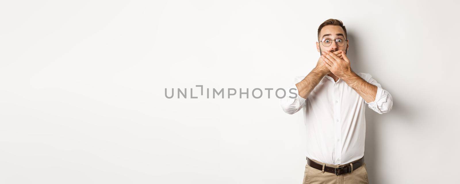 Shocked man gasping and looking at something in awe, covering mouth with hands, white background.