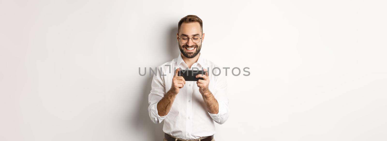 Happy businessman watching video on mobile phone, standing over white background.