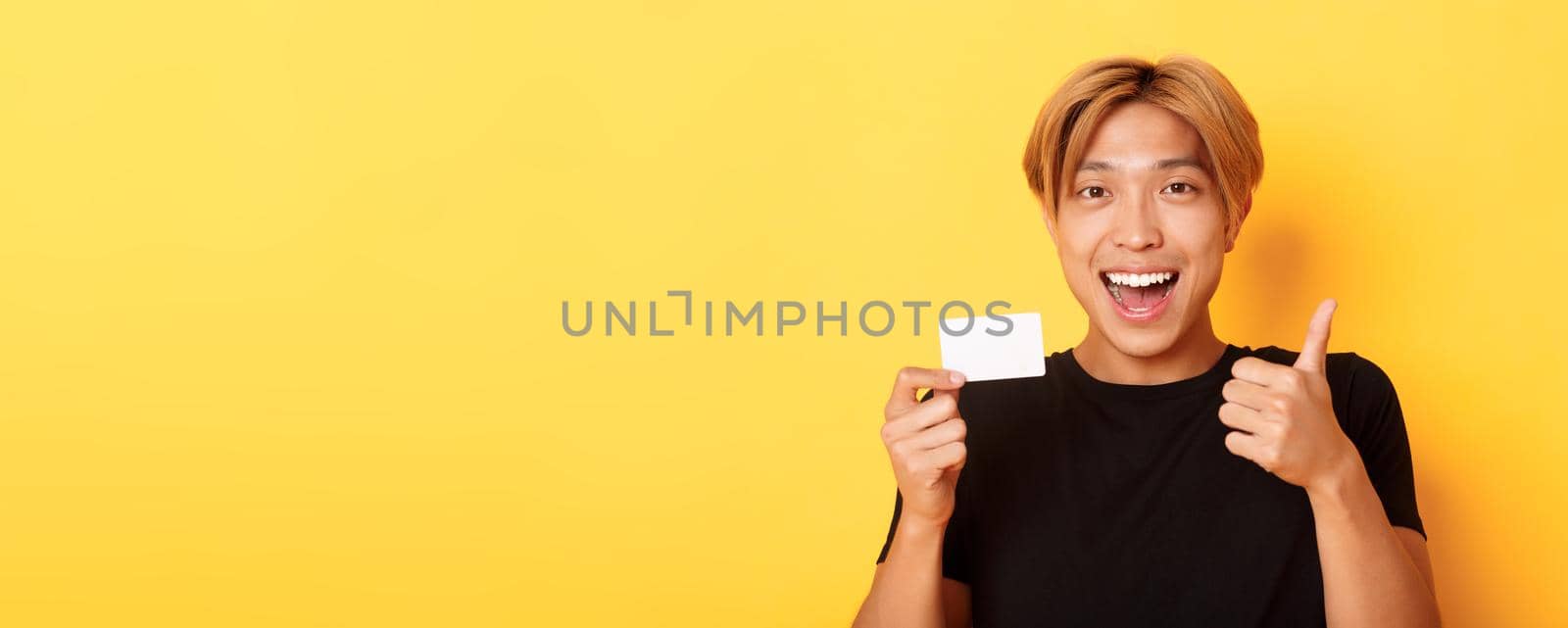 Close-up of happy and satisfied asian handsome guy, showing credit card and thumbs-up in approval, smiling amazed, standing yellow background.