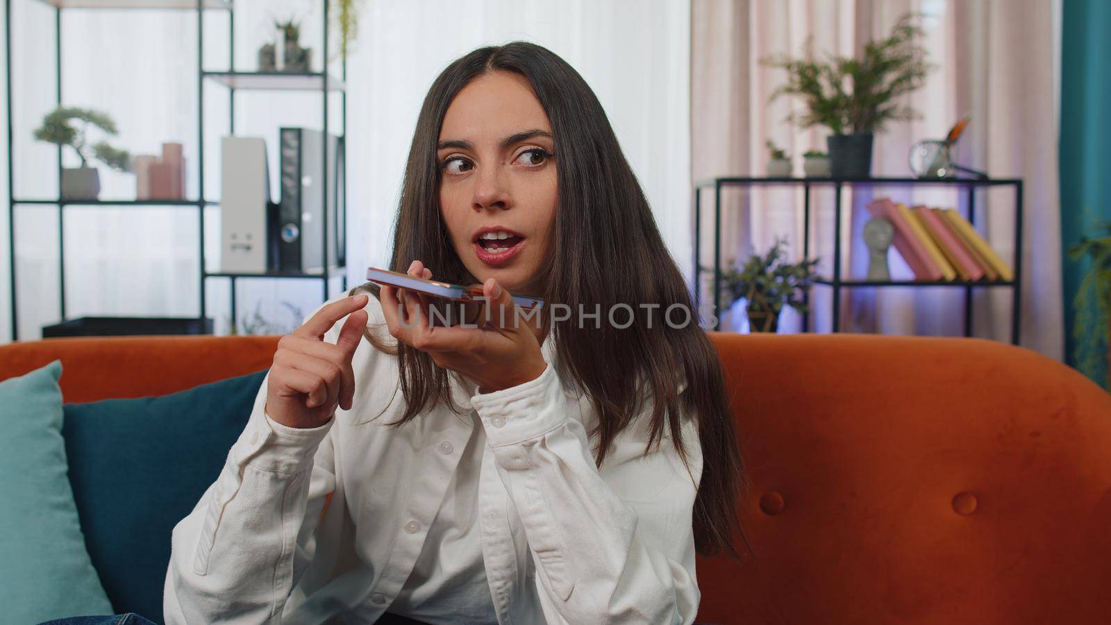 Portrait of caucasian girl making phone conversation with friends sitting call on couch at home in room. Happy excited young woman enjoying mobile loudspeaker talking indoors at modern home apartment