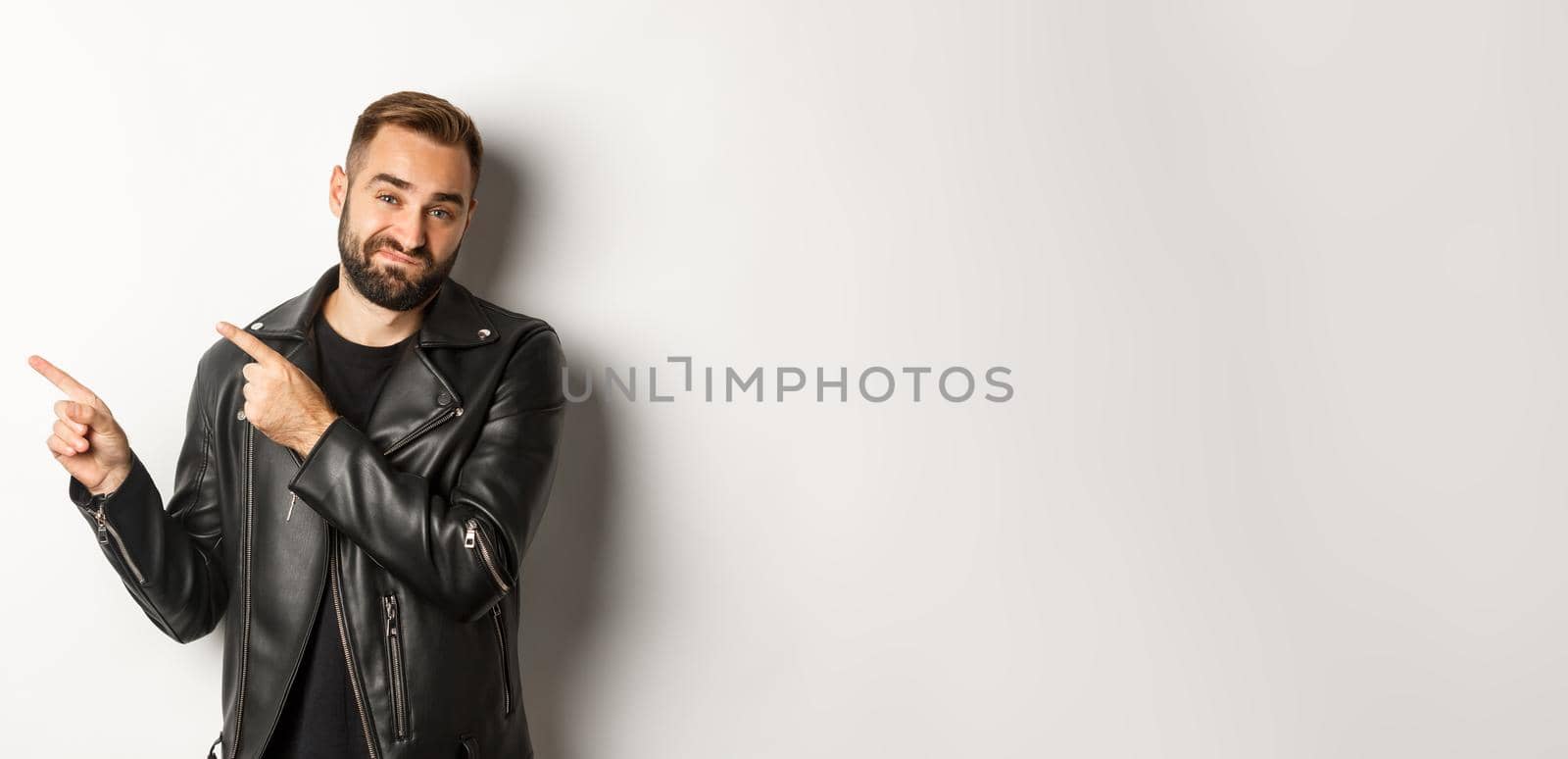 Skeptical and disapleased man in leather jacket, pointing fingers at upper left corner, showing bad promo offer, standing over white background reluctant.