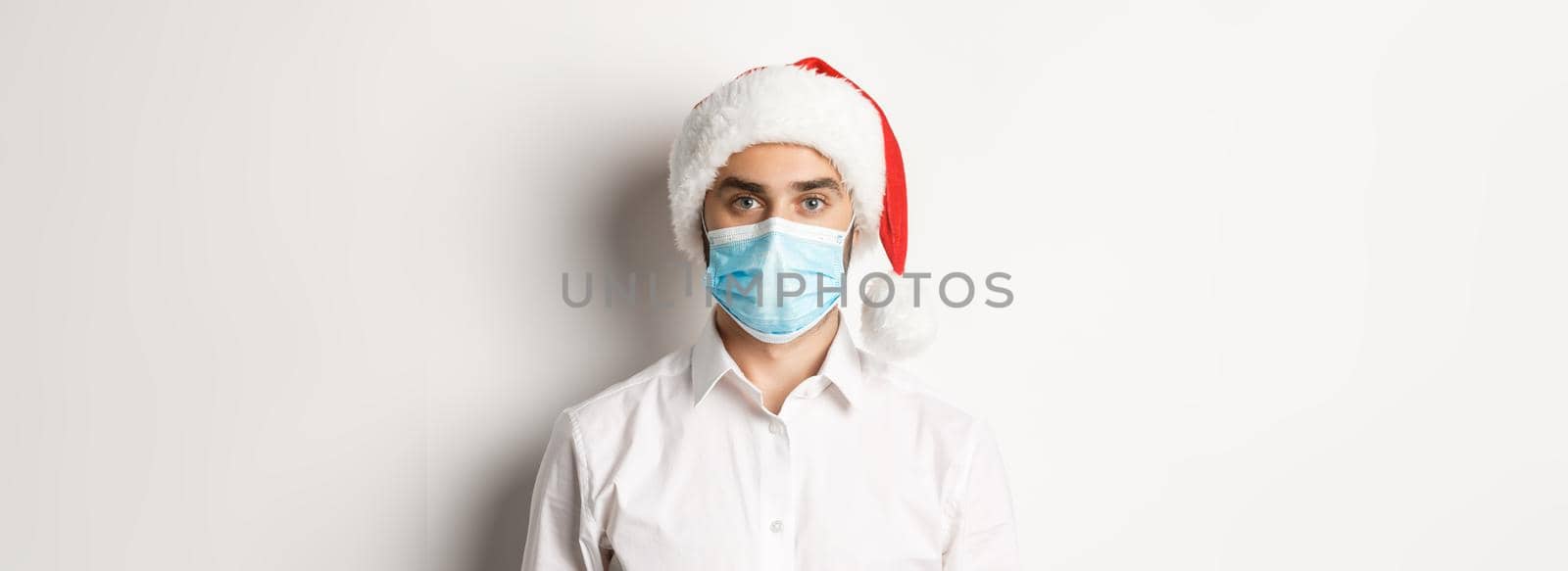 Concept of covid-19, social distancing and winter holidays. Close-up of young man wearing santa hat and face mask from coronavirus, celebrating New Year, white background.