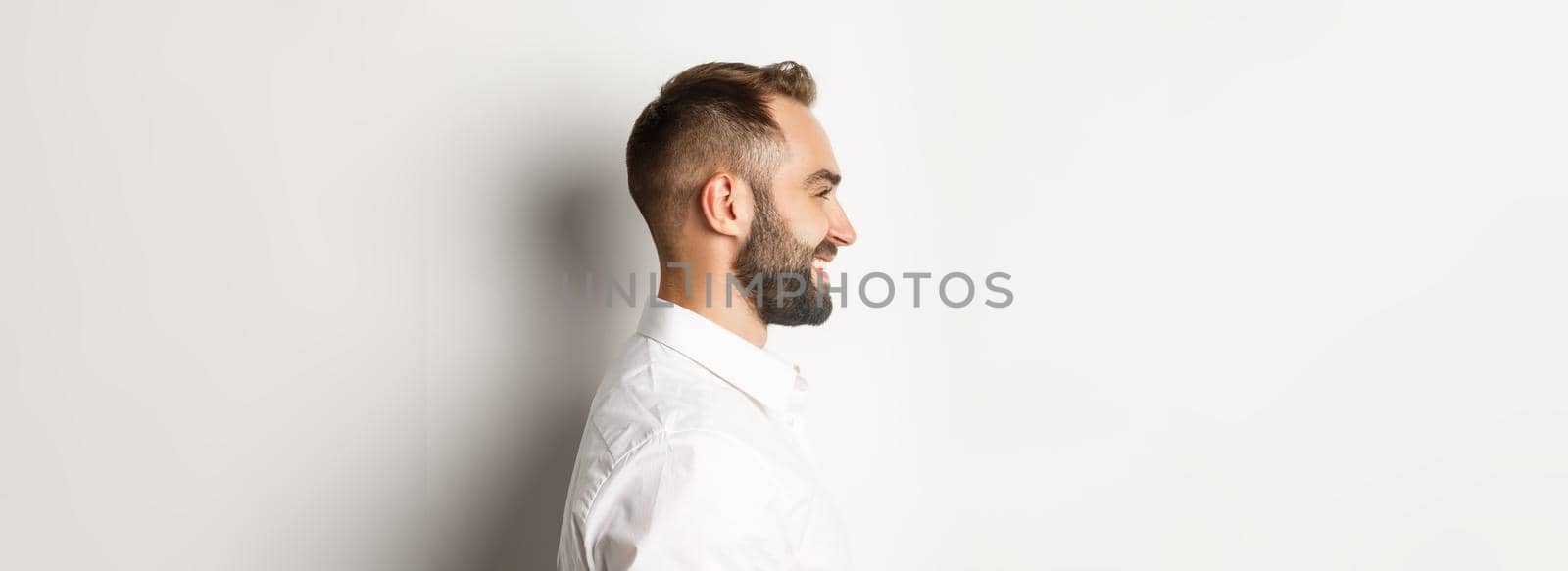 Close-up profile shot of handsome bearded man looking left and smiling, standing against white background.