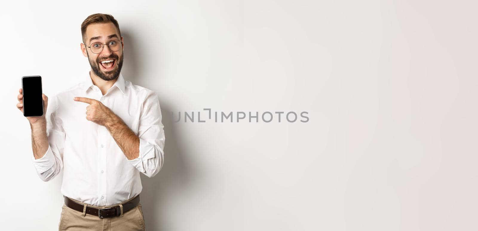 Excited handsome guy showing mobile phone, pointing finger at screen and smiling, standing against white background by Benzoix