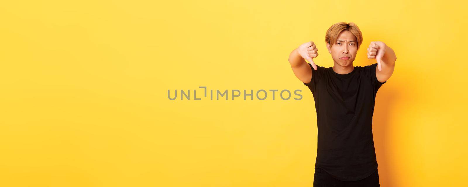 Portrait of disappointed and upset asian man showing thumbs-down, grimacing displeased, yellow background by Benzoix