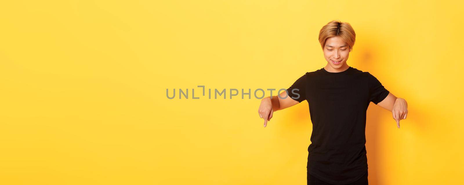 Portrait of curious happy blond asian guy, looking and pointing fingers down with pleased smile, yellow background.