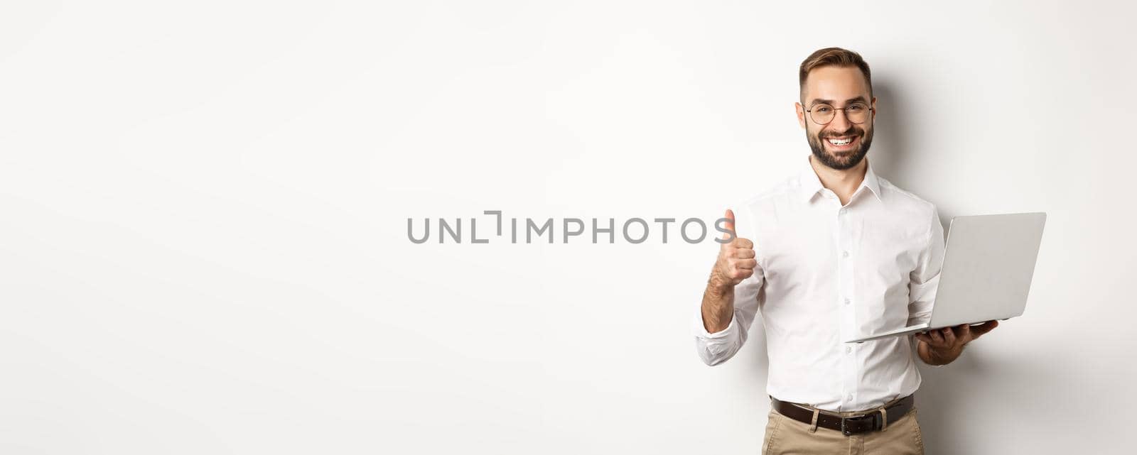 Business. Young successful male entrepreneur showing thumb-up while working on laptop, standing over white background.