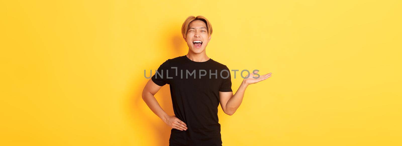 Portrait of happy blond asian guy, winking sassy and smiling, holding something on hand over yellow background.