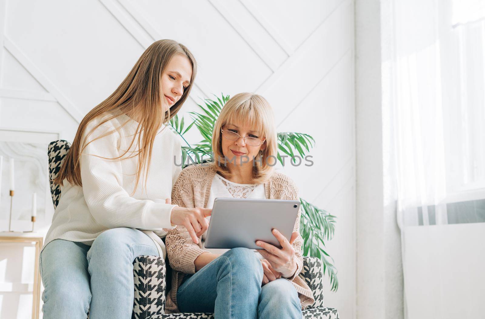 Senior woman and daughter playing with tablet pc. Smiling older woman and her granddaughter sitting home using gadget digital device for online shopping internet surfing video conference calling.