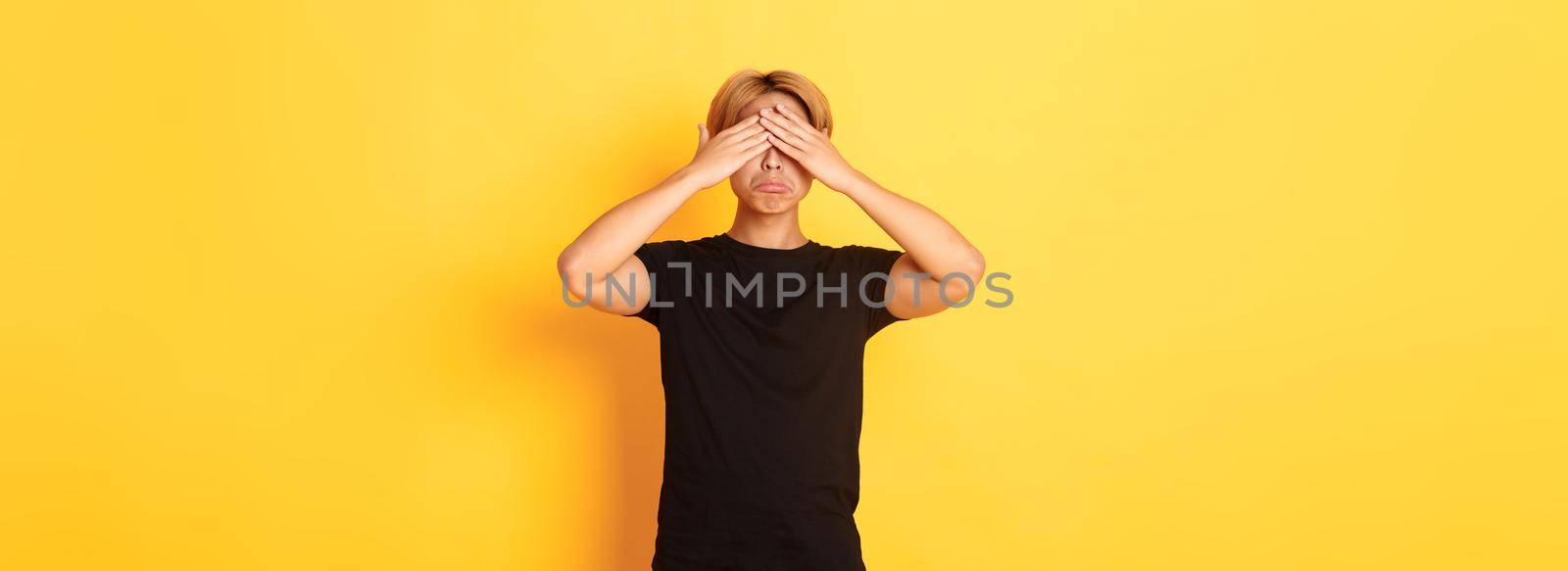 Portrait of sad and miserable asian guy shut eyes and sulking upset, standing yellow background.