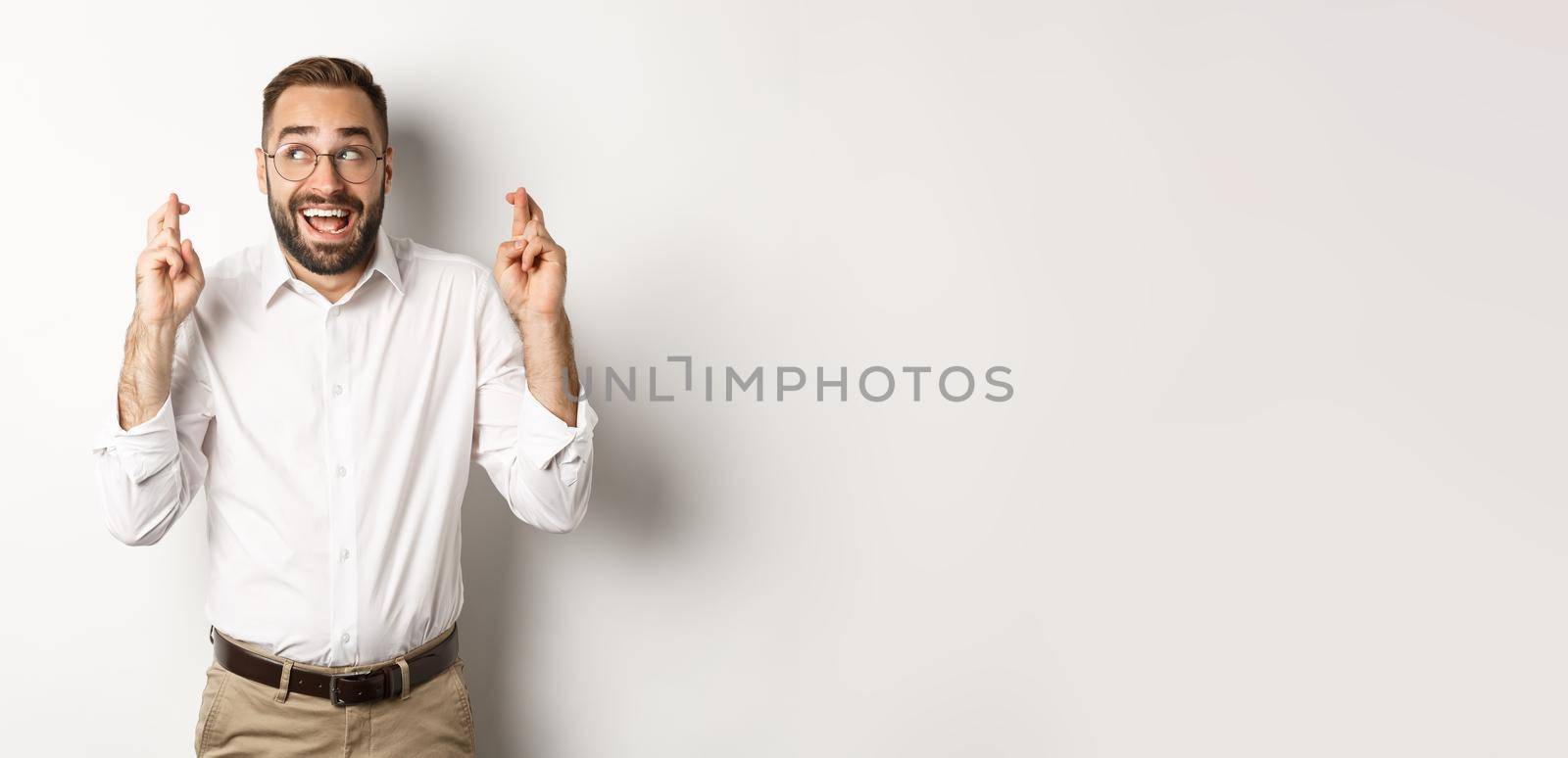 Excited and hopeful businessman making a wish, cross fingers and waiting, standing over white background.