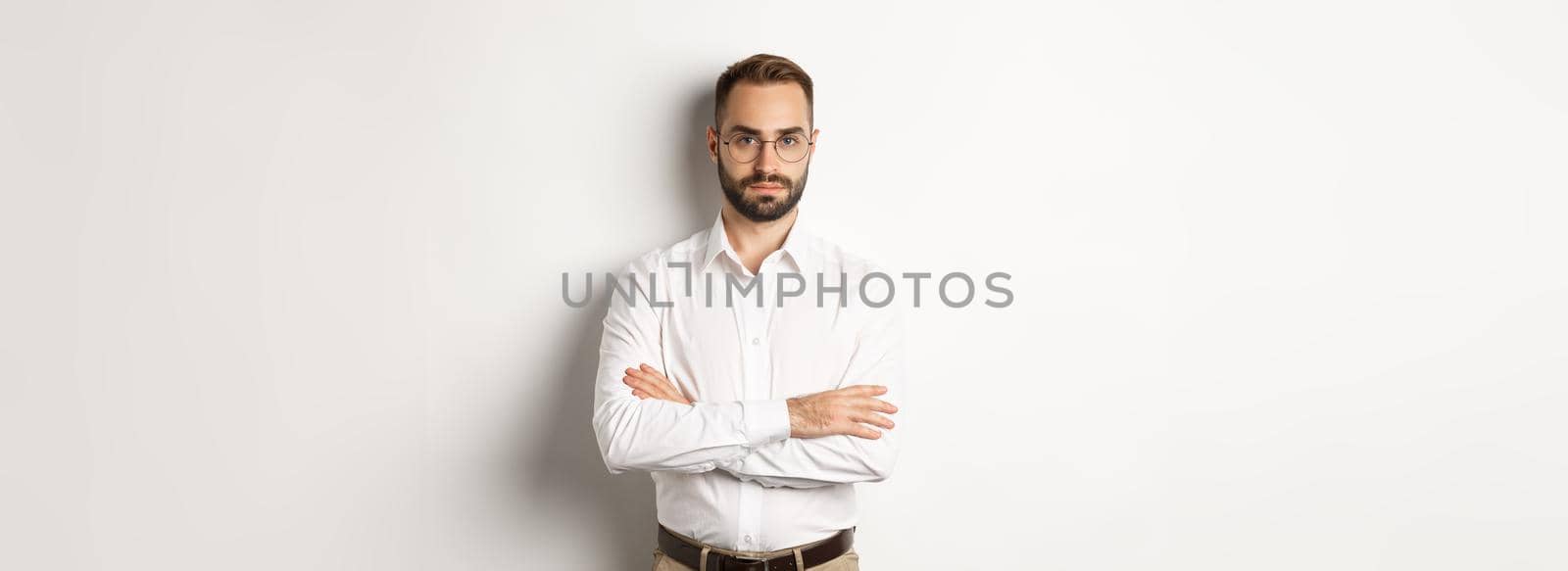 Confident businessman in glasses looking at camera, cross arms on chest, standing over white background.