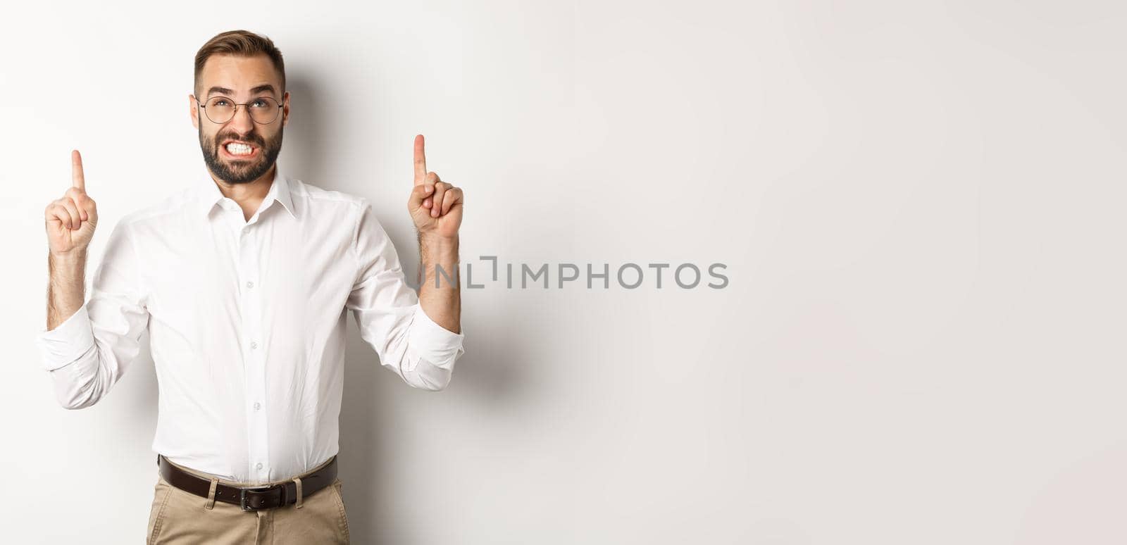 Doubtful and worried man looking and pointing fingers up, grimacing hesitant, standing over white background.