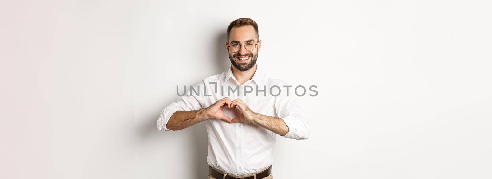 Handsome manage showing heart sign and smiling, I love you gesture, standing over white background.