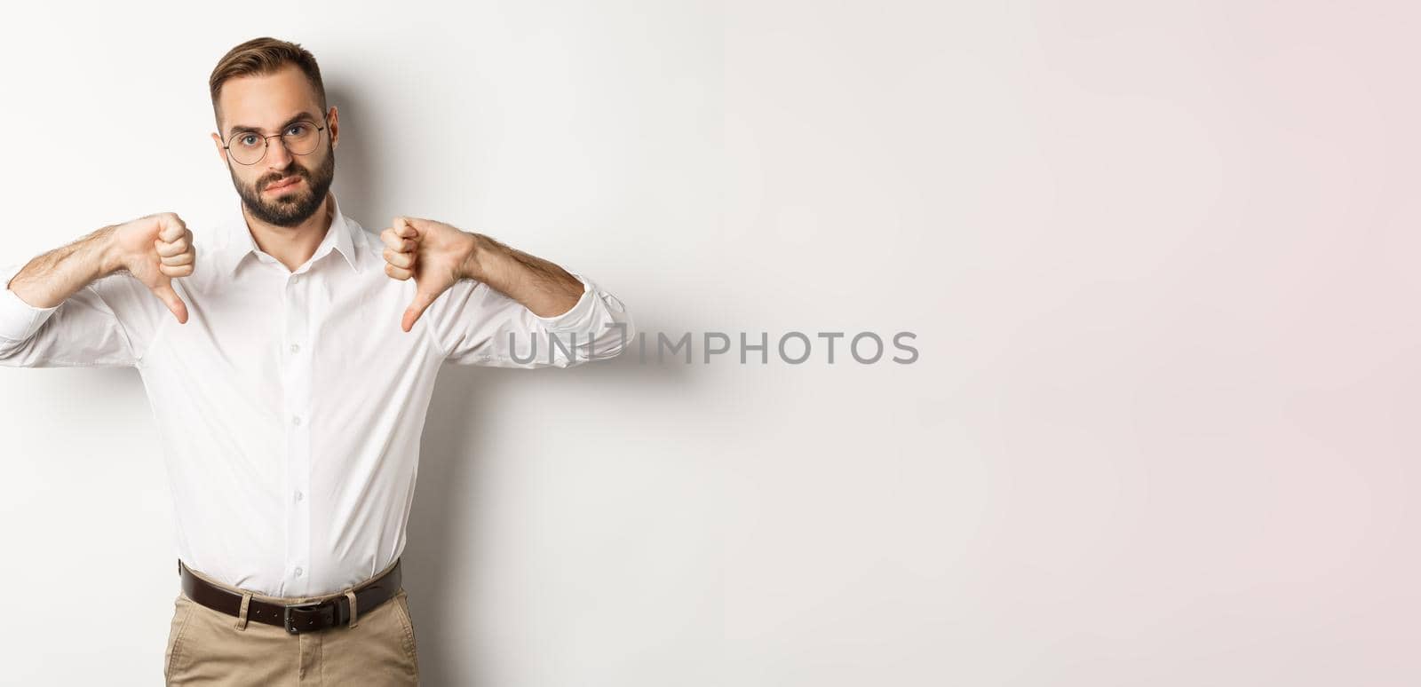 Displeased businessman in glasses showing thumbs down, dislike and disapprove, standing over white background.