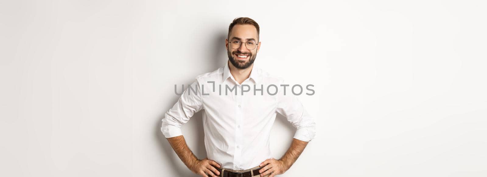 Handsome businessman in glasses looking satisfied, smiling and holding hands on waist, standing over white background.