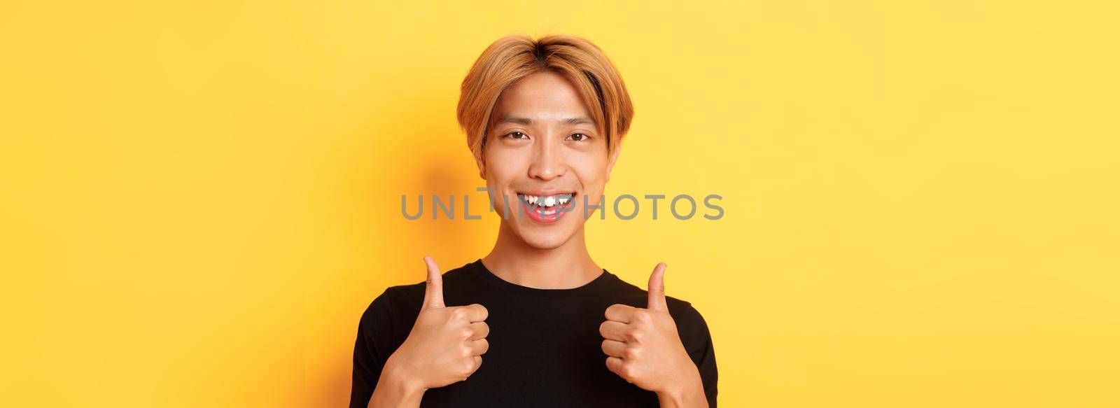 Close-up of satisfied, happy asian guy in black t-shirt, showing thumbs-up in approval, smiling and agree, standing yellow background by Benzoix