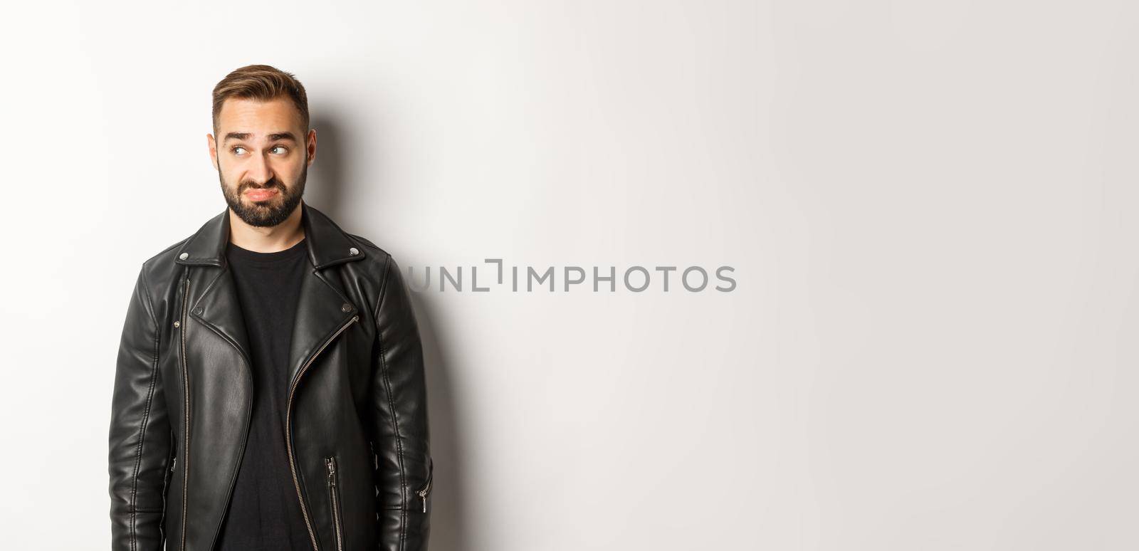 Indecisive gloomy man in black leather jacket, looking left and feeling uncomfortable, standing against white background.