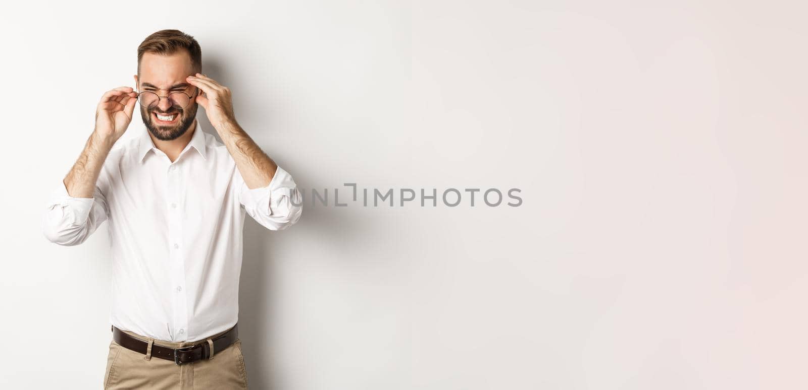 Businessman having headache, grimacing and holding hands on head, standing over white background.