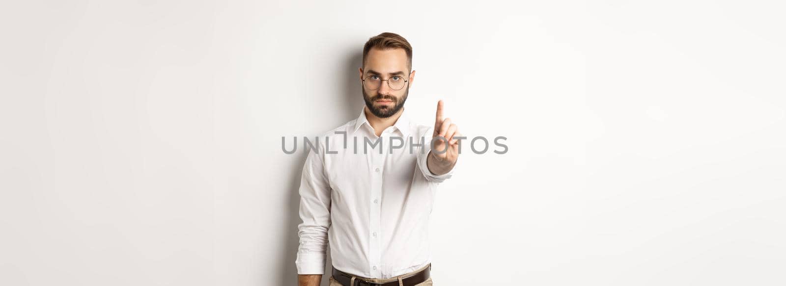 Serious man in glasses showing stop sign, shaking finger to prohibit and forbid, standing over white background.