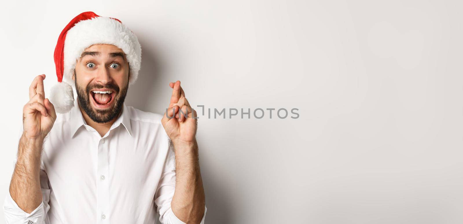 Party, winter holidays and celebration concept. Happy man in santa hat making christmas wish, cross fingers for good luck and looking excited, white background.