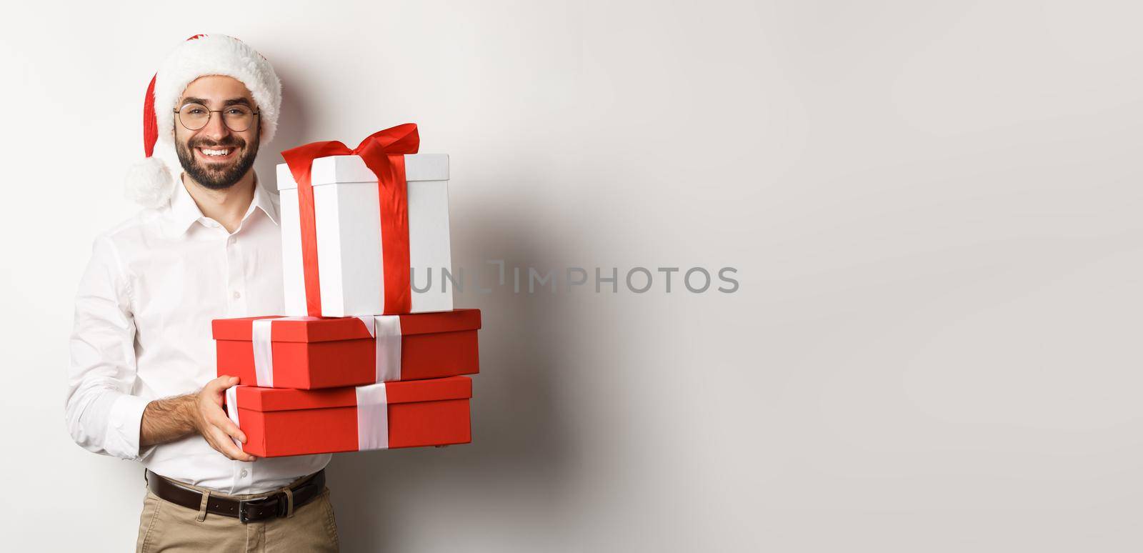 Merry christmas, holidays concept. Happy young man smiling, holding gifts in boxes and wearing santa hat, white background.