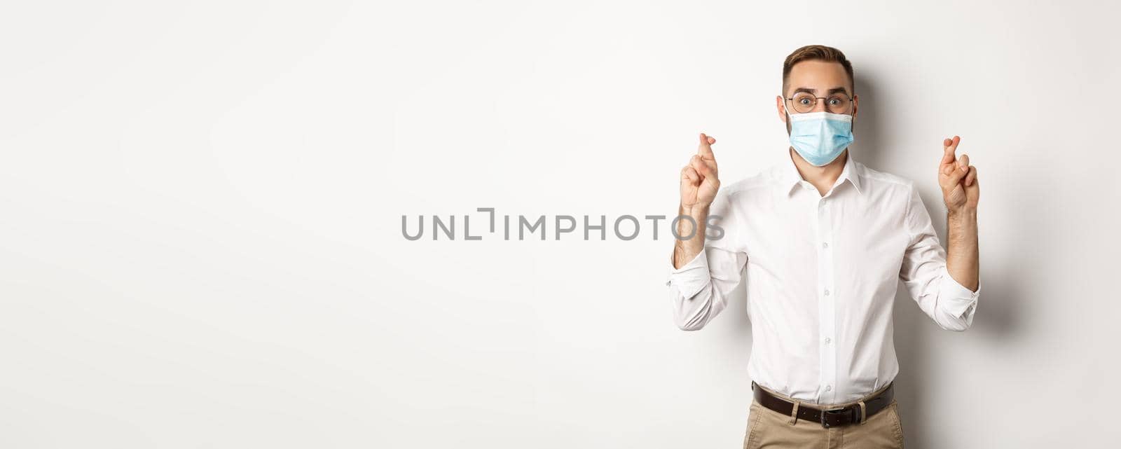 Covid-19, social distancing and quarantine concept. Hopeful employee in medical mask, holding fingers crossed and making wish, white background.