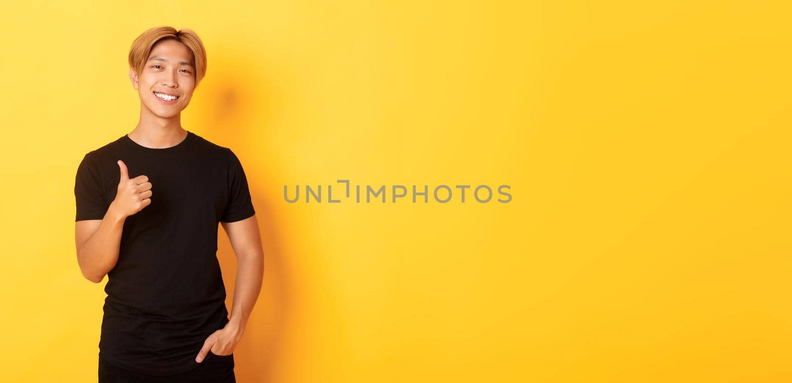 Portrait of satisfied handsome asian blond guy, smiling pleased, showing thumbs-up in approval, standing yellow background.