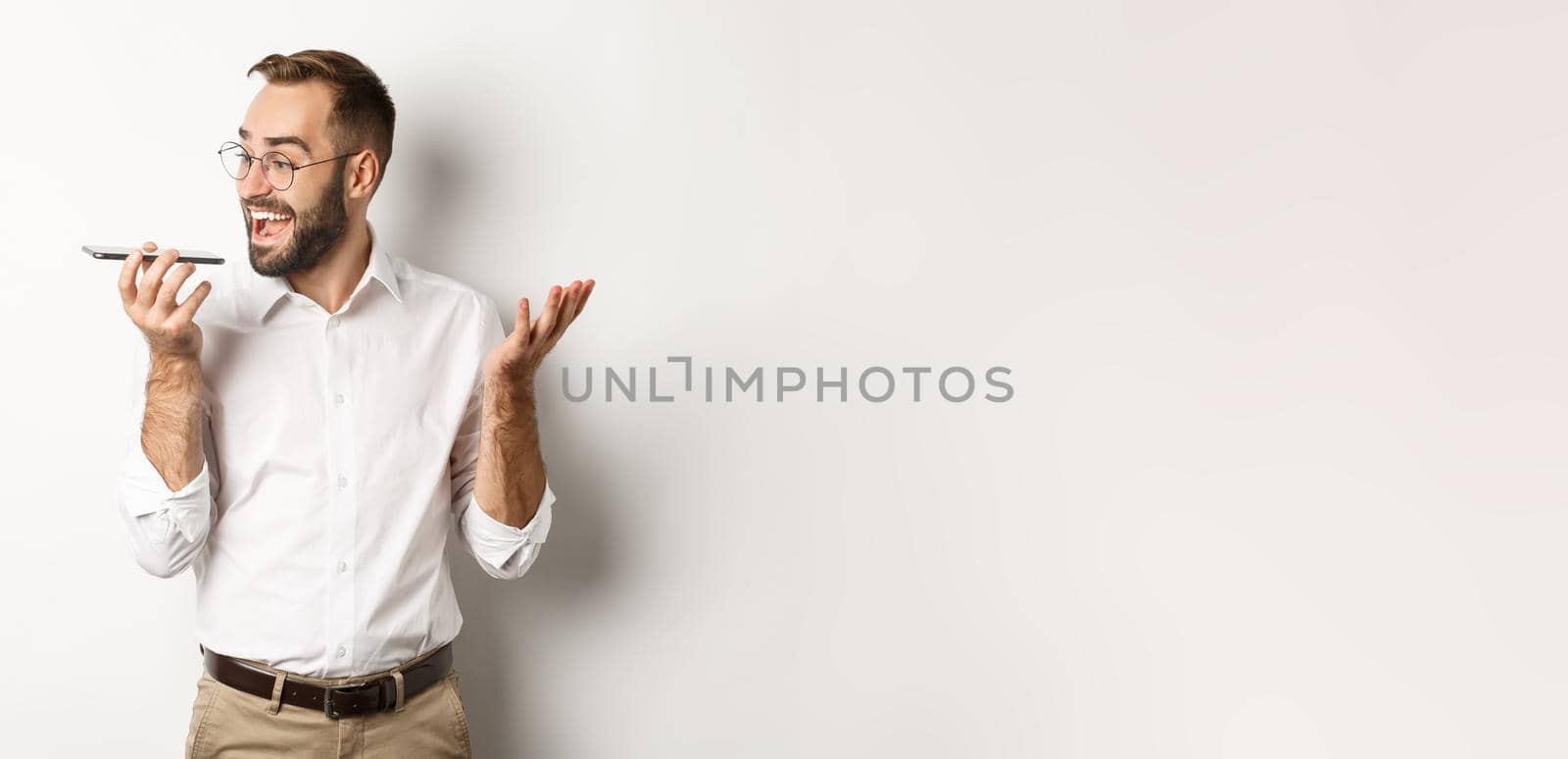 Excited businessman talking on speakerphone and smiling, record voice message with ecstatic face, standing over white background.