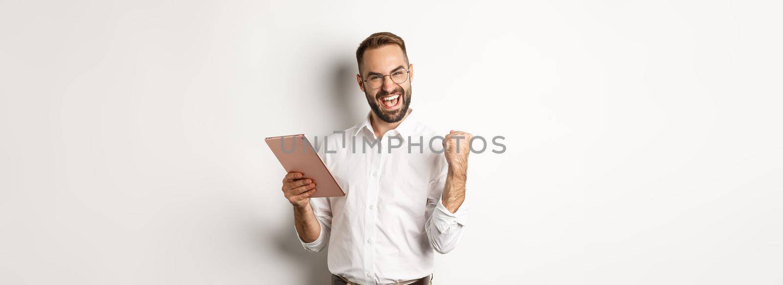 Successful businessman rejoicing on winning online, reading on digital tablet and making fist pump, triumphing, standing over white background.