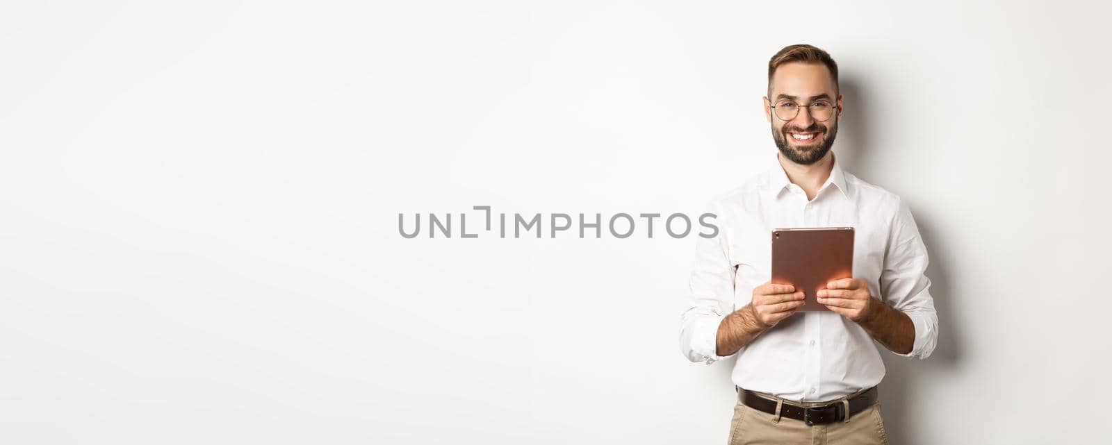Handsome bearded man in glasses using digital tablet, smiling satisfied, standing against white background.