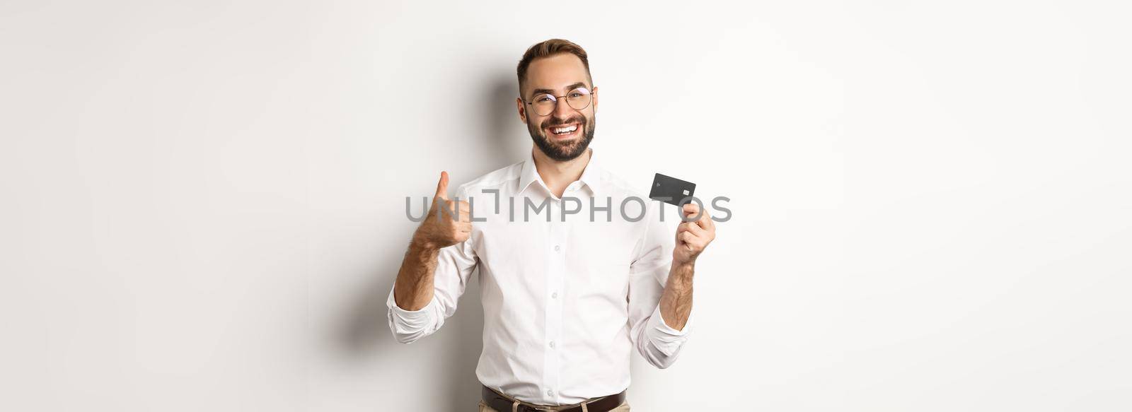 Handsome man showing his credit card and thumb up, recommending bank, standing over white background. Copy space