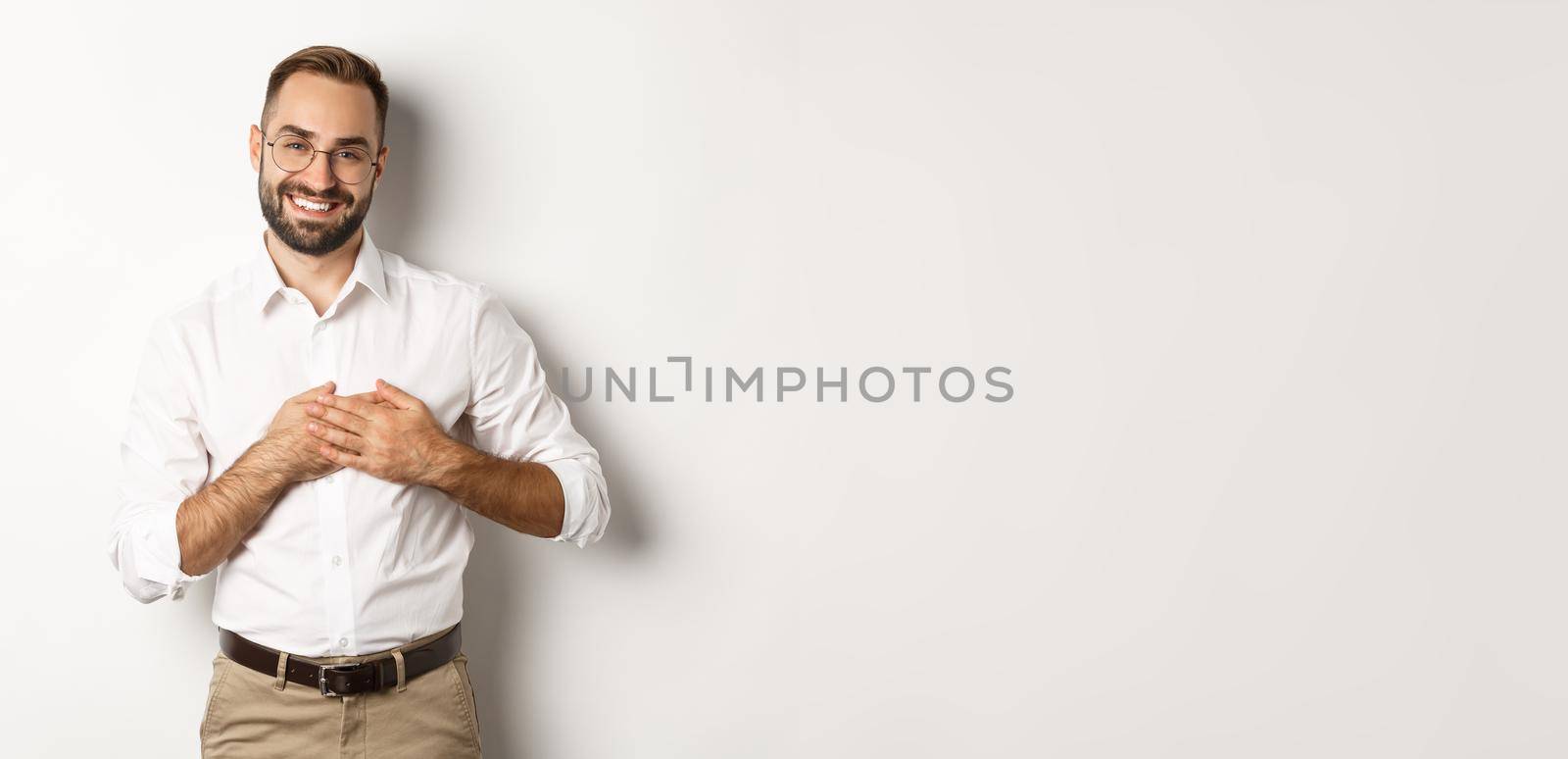 Touched and thankful business man holding hands on heart, smiling grateful, standing against white background.